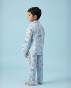 Back view of a young boy wearing light blue printed pajamas standing barefoot against a pastel blue background.