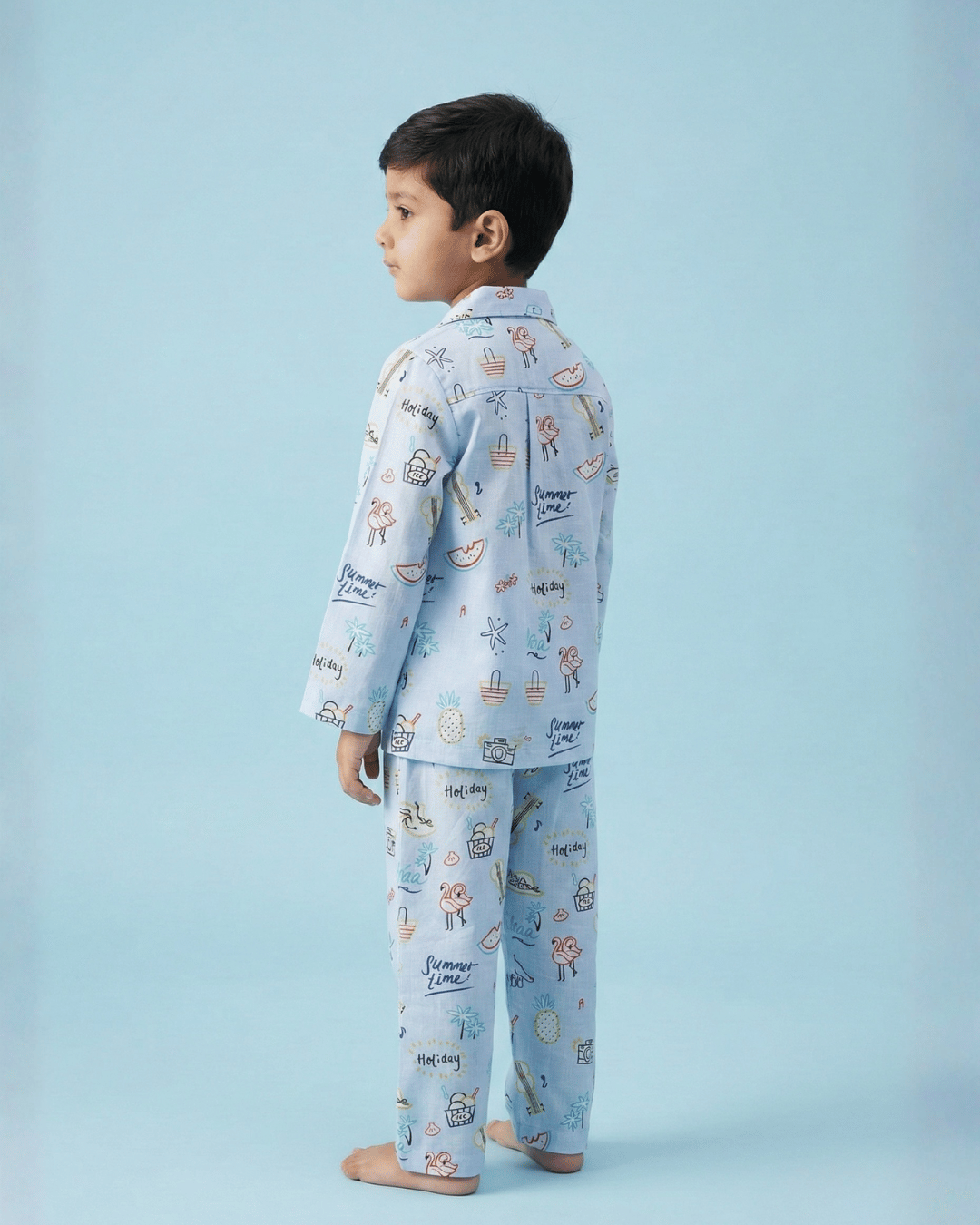 Back view of a young boy wearing light blue printed pajamas standing barefoot against a pastel blue background.