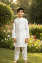 Young boy in a white traditional outfit standing in a garden with greenery and flowers.